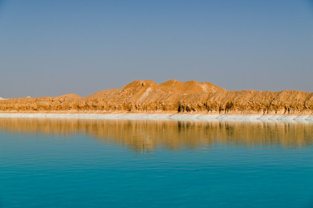 Turquoise water reflects a sandy, rocky shoreline under blue sky.